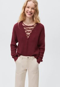 Burgundska pletena jopica z globokim V-izrezom in križnim vezanjem, v kombinaciji s kremnimi ravno krojenimi hlačami.