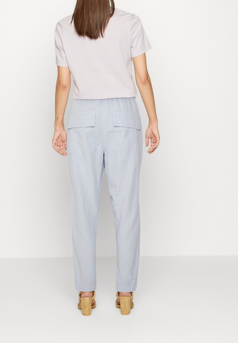 Femme portant un pantalon gris clair avec poches arrière, un haut blanc à manches courtes et des sandales à talons beiges, debout de dos sur un fond uni.