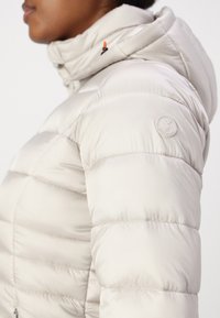 Giacca puffer beige chiaro con cappuccio, con texture trapuntata, un logo circolare sulla spalla e un accento arancione sul cordino del cappuccio.