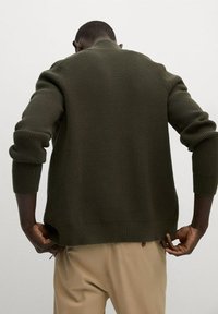 Jersey de canalé verde oscuro con cuello alto y mangas largas, que presenta aberturas laterales en el dobladillo para facilitar el movimiento.