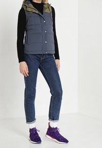 Vadderad väst i mörkgrått med en olivgrön huva, ihop med mörkblå jeans och lila sneakers. Vesten har knappar och fickor.