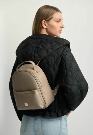 Młoda kobieta w czarnej pikowanej kurtce i jasnoniebieskich dżinsach niesie na plecach beżowy skórzany plecak, patrząc na bok.