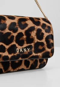 Kabelka s leopardím vzorem, se zlatým kováním, řetízkem a měkkou kožešinovou texturou, s logem DKNY na přední straně. Hlavní barvy jsou béžová a černá.
