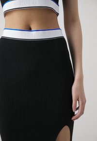 Femme portant une jupe noire côtelée et un top court assorti avec des bandes élastiques blanches à la taille, arborant un logo discret et une bordure bleue.
