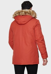 Parka orange isolée avec une capuche en fausse fourrure, présentant une texture lisse, des manches longues et des poches latérales pour une fonctionnalité accrue.
