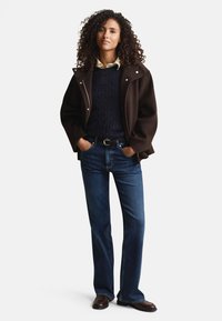 Veste en mélange de laine marron avec grand col, pull marin en tricot torsadé bleu marine, chemise jaune clair, jean bleu à jambes droites, et chaussures en cuir marron.