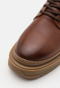 Brązowy skórzany but z zaokrąglonym noskiem, z grubą beżową podeszwą i teksturowanymi detalami wokół sznurowadeł oraz górnej krawędzi.
