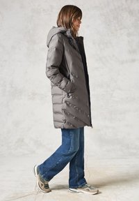 Abrigo acolchado gris con diseño de costuras horizontales, capucha y detalles en azul marino; combinado con vaqueros acampanados azules y zapatillas grises.