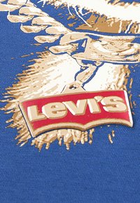 Μπλε ύφασμα με γραφικό σχέδιο σε χρυσό και λευκό που απεικονίζει ένα φύλλο δάφνης και μια κόκκινη ετικέτα Levi's με λευκό κείμενο, περιγραμματισμένη με χρυσό.