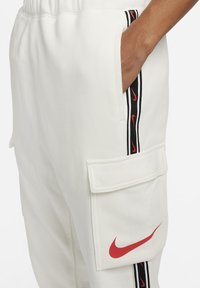 White sweatpants with an elastic waistband, side pocket, and a cargo pocket featuring a red Nike logo and black side strips with red accents.