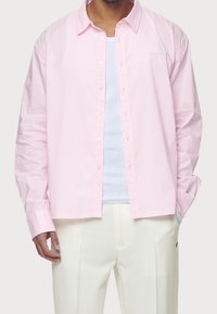 Jasnoróżowa, w paski, koszula zapinana na guziki z klasycznym kołnierzykiem i długim rękawem, z białymi guzikami i luźnym krojem. Nosi się ją na białej bluzce.