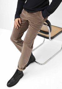 Bruine corduroy broek met verticale ribbels, gecombineerd met zwarte leren veterschoenen. Een zwart langemouwen top is gedeeltelijk zichtbaar.