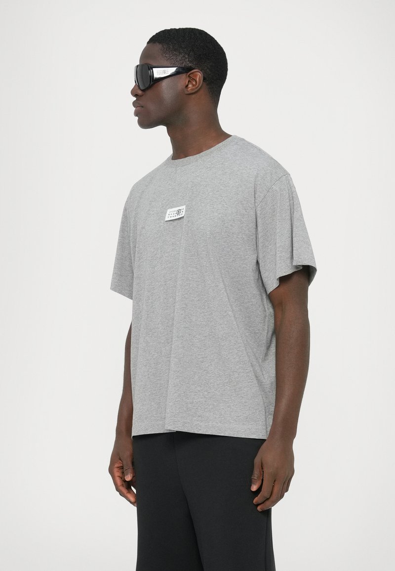 Gray cotton t-shirt with short sleeves and a relaxed fit, featuring a small graphic label on the chest and paired with black sunglasses.