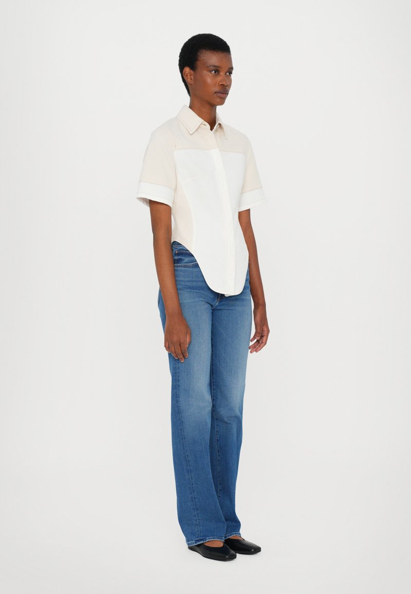 Short-sleeve shirt in beige with a white panel, paired with blue flared jeans. The outfit features a collared neckline and soft fabric texture.