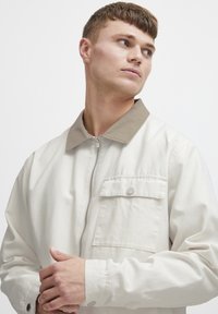 Lichtbeige jas met een contrasterende tan kraag, ritssluiting en een voorzak. De stof lijkt glad met een casual ontwerp.