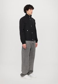 Black lightweight jacket with high collar, front snap buttons, and two chest pockets. Paired with gray wide-leg jeans and white sneakers.