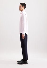 Lichtroze langemouw shirt, van katoen, met een kraag. Draagt een marineblauwe broek en zwarte leren schoenen, staand tegen een neutrale achtergrond.