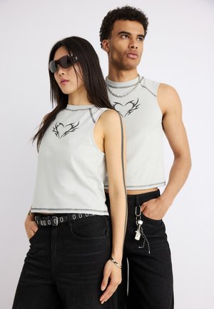 Δύο μοντέλα που φορούν ταιριαστά χωρίς μανίκια λευκά τοπ με μαύρα τατουάζ καρδιάς, μαύρα παντελόνια, το ένα με γυαλιά ηλίου και ζώνη, το άλλο με κολιέ αλυσίδα.