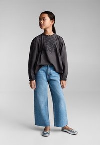 Bluză lungă gri cu mâneci lungi, brodată cu flori, purtată cu blugi largi albaștri și balerini argintii. Design simplu și casual.