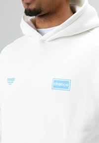 Witte hoodie met een zachte textuur. Heeft blauwe tekstpatches: "STRONGER" links en "chance" rechts, beide in rechthoekige ontwerpen.