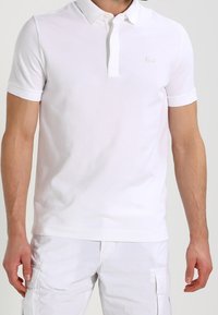 Polo shirt blanc à manches courtes en coton. Il présente un col plat, une patte de boutonnage à trois boutons, un logo subtil sur la poitrine et une texture lisse.