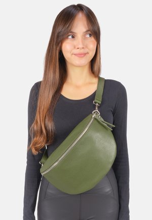 Junge Frau trägt ein schwarzes Oberteil und schwarze Hose mit einer großen grünen Umhängetasche schräg über der Brust, lächelnd und seitlich schauend.