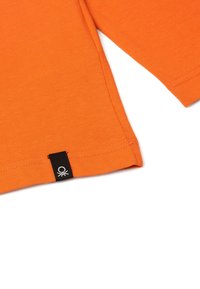 Oranžové tričko s krátkým rukávem, které má žebrovanou strukturu, rovný střih a malou černou nášivku s logem na spodním lemu.