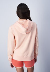 Sudadera color durazno con capucha ajustable, mangas largas y una textura suave, combinada con shorts naranjas brillantes con dobladillo en ondas.