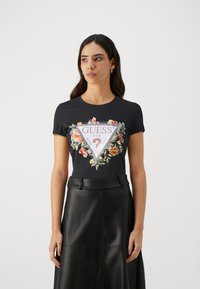 T-shirt noir avec un design floral et le logo "GUESS" dans un motif triangulaire. Le t-shirt a des manches courtes et est associé à une jupe en cuir noir.