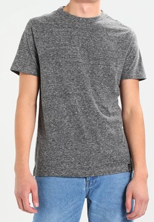 Ung person iført en kortærmet gråmeleret t-shirt og blå jeans, stående med armene afslappet ned langs siderne.
