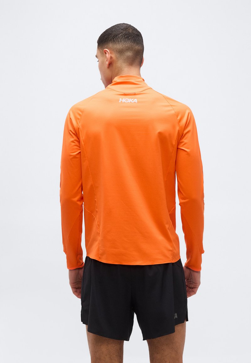 Mann trägt ein hellorangefarbenes, langärmeliges Sportoberteil und schwarze, sportliche Shorts, steht mit dem Rücken zur Kamera vor einem einfarbigen hellen Hintergrund.