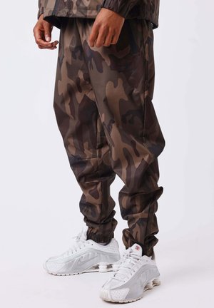 Personne portant un pantalon camouflage marron et vert foncé et des baskets blanches Nike Shox, debout devant un fond uni.