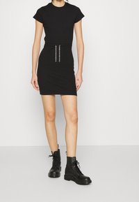 Robe noire moulante à manches courtes, avec un détail en chaîne à la taille. Assortie à des bottes de combat noires à lacets. Texture lisse.