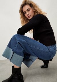 Pantaloni a gamba larga in denim blu con risvolti azzurri a contrasto, orli sfrangiati e vita alta, abbinati a stivaletti neri alla caviglia.