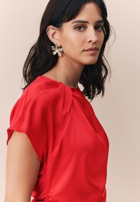 Červená krátká halenka s krátkým rukávem a zdobením na ramenech. Modelka nosí zlaté náušnice ve tvaru květin. Hladká textura, přiléhavý střih.