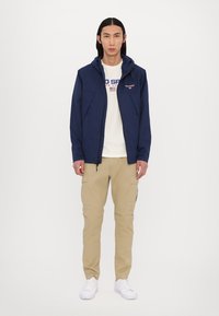 Marineblå zip-up jakke med hætte, lavet af blandet stof, parret med en hvid grafisk T-shirt og beige cargo bukser. Hvide sneakers fuldender looket.