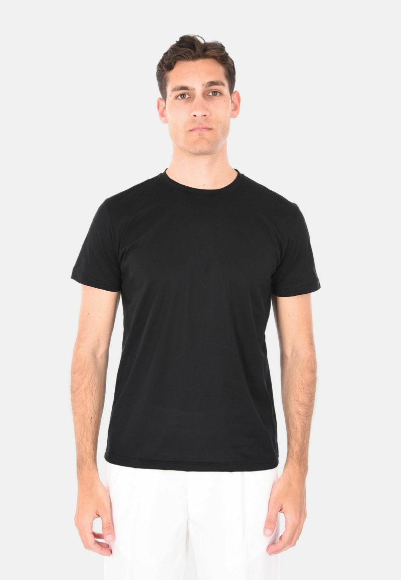 T-shirt nera in cotone con scollo rotondo, maniche corte e una texture liscia. Indossata con pantaloni bianchi, esposta su uno sfondo neutro.