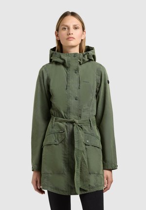 Donna che indossa una giacca con cappuccio verde con cintura e grandi tasche frontali, in piedi davanti a uno sfondo grigio uniforme.