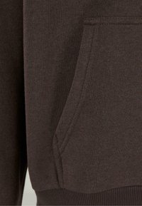 Tejido de sudadera marrón con una textura suave, que cuenta con un bolsillo lateral y puños acanalados. Se puede ver el detalle de las costuras cerca del bolsillo.