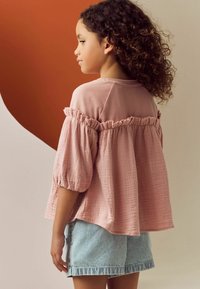 Hellrosa, Oberteil mit Rüsche und aus leichtem Stoff, kombiniert mit blauen und weißen gestreiften Jeansshorts, von hinten gezeigt.