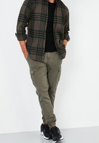 Man med uppvikta ärmar i rutig skjorta, svart t-shirt, olivgröna cargo-byxor och svarta sneakers, står med händerna i fickorna.