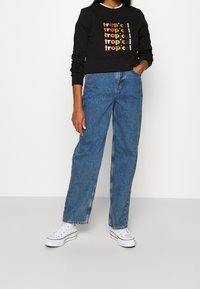 Mujer vestida con un sudadera negra con texto colorido "tropical", jeans de tiro alto azules y zapatillas de plataforma blancas, de pie con una mano en el bolsillo.