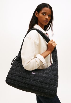 Mujer con cabello largo y oscuro que lleva una chaqueta ligera y un gran bolso de hombro acolchado negro de Tommy Jeans.