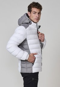 Giacca imbottita bianca e grigia, con cappuccio, pannelli a contrasto e tasche frontali con zip. Il tessuto ha una texture liscia.