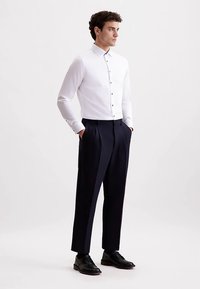 Camicia bianca a maniche lunghe con chiusura frontale a bottoni, abbinata a pantaloni blu scuro con pieghe, indossata con scarpe nere.