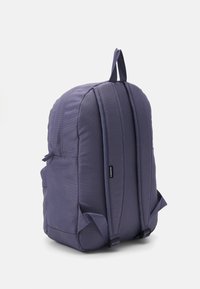 Mochila de tela morada con correas de hombro acolchadas, asa en la parte superior y pequeño bolsillo lateral, mostrada desde atrás sobre un fondo blanco.