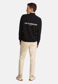 Felpa nera con la scritta "LES CLASSIQUES" in bianco sulla schiena, abbinata a pantaloni beige chiaro e sneakers beige. Design moderno, stile casual.