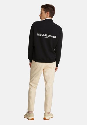 Felpa nera con la scritta "LES CLASSIQUES" in bianco sulla schiena, abbinata a pantaloni beige chiaro e sneakers beige. Design moderno, stile casual.