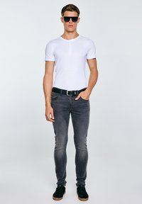 Mann mit schwarzen Sonnenbrillen, weißem kurzärmeligem Hemd, schwarzem Gürtel, grauen Skinny-Jeans und schwarzen Turnschuhen, der mit einer Hand in der Tasche steht.