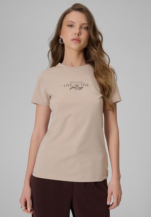 Młoda kobieta z długimi falującymi włosami, ubrana w beżową koszulkę z napisem „Live Active feel cozy” oraz brązowe spodnie na szarym tle.